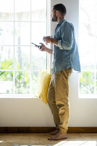 Junger Mann zu Hause, der sein Smartphone und seinen Kaffeebecher in der Hand hält und nach draußen schaut, lizenzfreies Stockfoto