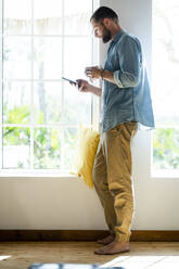 Young man at home standing at window and checking his smartphone - SBOF02275