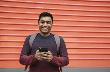 Porträt eines lächelnden Mannes, der ein Smartphone mit rotem Hintergrund hält - AHSF02094