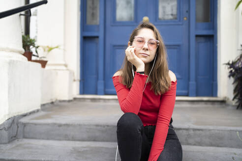 Porträt einer jungen Frau, die auf einer Treppe am Hauseingang sitzt und Kopfhörer trägt - FBAF01349