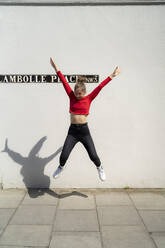 Exuberant young woman jumping in front of a wall - FBAF01357