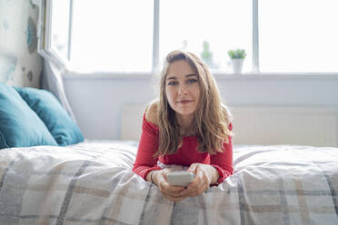 Portrait of young woman lying on bed at home holding cell phone - FBAF01371