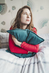 Ernste junge Frau sitzt zu Hause im Bett und umarmt ein Kissen - FBAF01372