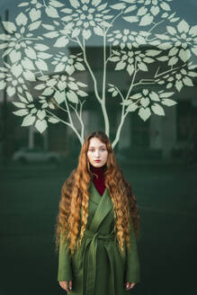 Portrait of redheaded young woman wearing green coat standing in front of glass pane with stylised tree - TCEF00335