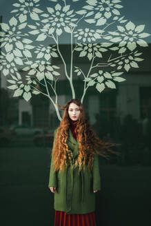 Portrait of redheaded young woman wearing green coat standing in front of glass pane with stylised tree - TCEF00336