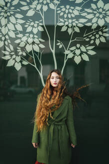 Porträt einer rothaarigen jungen Frau in grünem Mantel vor einer Glasscheibe mit stilisiertem Baum - TCEF00337