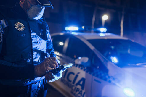 Policeman during emergency mission at night, taking notes, wearing protective gloves and mask - JCMF00524