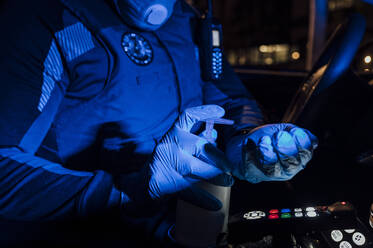 Polizist mit Maske, Schutzhandschuhen und Desinfektionsmittel während eines nächtlichen Noteinsatzes - JCMF00530