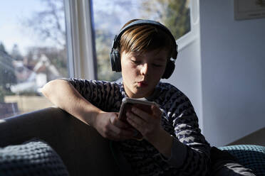 Porträt eines pfeifenden Jungen, der mit Kopfhörern Musik hört, während er auf sein Mobiltelefon schaut - AUF00250