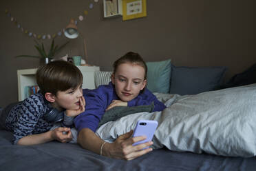 Portrait of brother and his older sister lying together on bed using smartphone for video chat - AUF00253