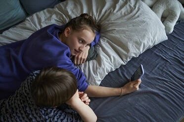 Geschwister liegen zusammen auf dem Bett und benutzen ihr Smartphone für einen Videochat - AUF00254