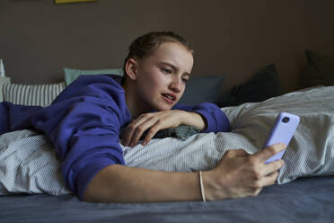 Portrait of girl lying on bed at home using smartphone for video chat - AUF00257