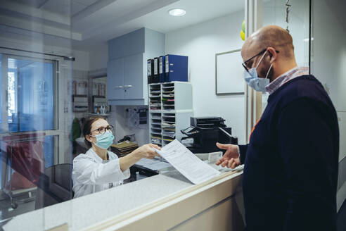 Employee at reception desk of hospital ward handing over application form to visitor - MFF05291