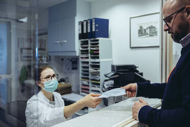 Employee at reception desk of hospital ward handing over mask to visitor - MFF05292