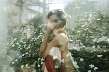 Portrait of young woman standing behind glass pane in an urban garden - TCEF00370