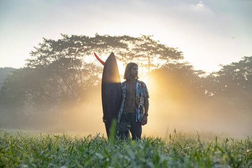 Junger Mann mit Surfbrett auf einer Wiese bei Sonnenaufgang, Costa Rica - AMUF00027