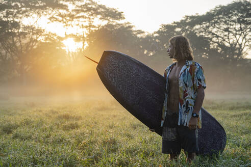 Junger Mann mit Surfbrett auf einer Wiese bei Sonnenaufgang, Costa Rica - AMUF00029