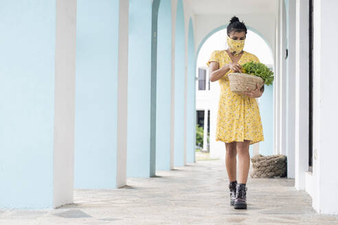 Woman wearing mouth cloth holding basket with salad and fresh fruits - AMUF00034