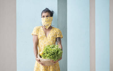 Woman wearing mouth cloth holding basket with salad and fresh fruits - AMUF00036