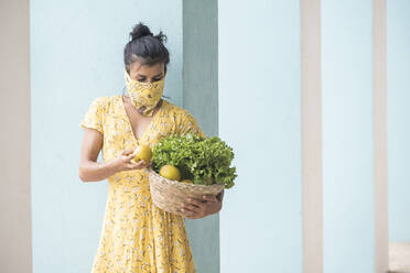 Woman wearing mouth cloth holding basket with salad and fresh fruits - AMUF00037