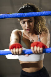 Athletic female boxer in boxing ring leaning on rope concentrating before the fight - JSMF01498