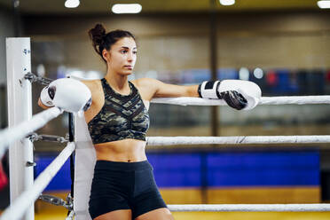 Athletic female boxer in boxing ring leaning on rope in the corner - JSMF01530