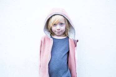 Portrait of little blond girl wearing pink hooded jacket in front of white wall - IHF00309