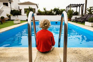 Back view of little boy sitting in front of a swimming pool - IHF00314