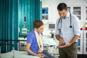 Male pediatrician showing digital tablet to boy patient with arm in sling in hospital - CAIF24912