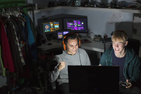 Aufgeregte Teenager mit Kopfhörern spielen ein Videospiel am Computer in einem dunklen Raum, lizenzfreies Stockfoto