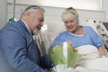 Älterer Mann bringt seiner im Krankenhausbett liegenden Frau einen Blumenstrauß - CAIF25063
