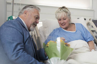 Senior man bringing flower bouquet to wife resting in hospital bed - CAIF25063