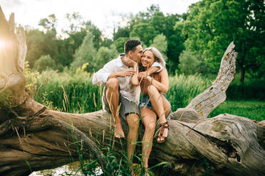 Junges Paar sitzt auf einem umgestürzten Baum am Flussufer, lächelt und umarmt sich. - ISF23981