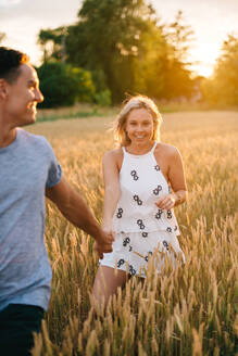 Ein lächelndes junges Paar geht Hand in Hand durch ein goldenes Weizenfeld. - ISF23985