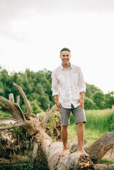 Young man wearing shorts standing on fallen tree on a riverbank, smiling at camera. - ISF23987