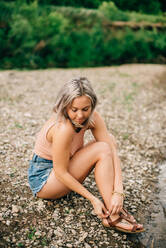 Junge blonde Frau in kurzen Hosen sitzt am Flussufer und zieht Sandalen an. - ISF23993