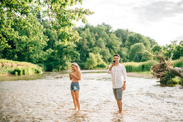 Ein junges Paar steht knöcheltief in einem Fluss und fotografiert mit dem Handy. - ISF23996