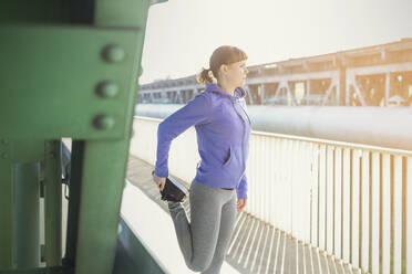 Young female runner stretching leg along sunny urban railing - CAIF25148