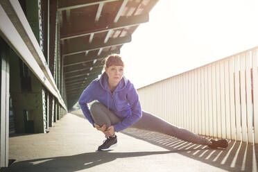 Focused young female runner stretching legs on sunny train station platform - CAIF25158