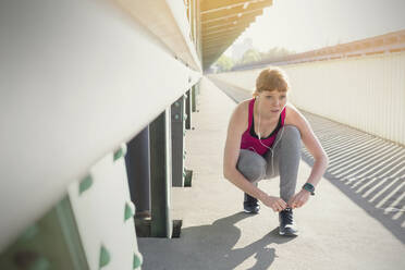 Focused young female runner tying shoe on sunny train station platform - CAIF25174