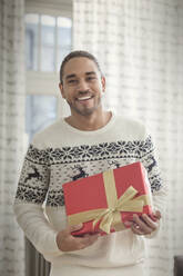 Portrait smiling young man in Christmas sweater holding gift - CAIF25201