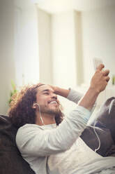 Happy young man listening to music with headphones and mp3 player on sofa - CAIF25203