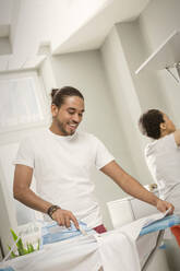 Young man doing laundry, ironing shirt in laundry room - CAIF25213
