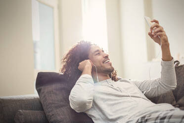 Lächelnder junger Mann, der sich mit Smartphone und Kopfhörern auf dem Sofa entspannt und Musik hört - CAIF25218