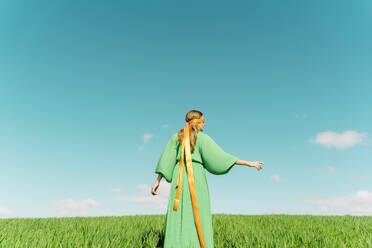 Rückansicht einer jungen Frau in einem grünen Kleid, die mit einer Schleife um den Kopf auf einem Feld steht - ERRF02997