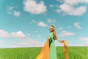 Happy young woman wearing a green dress in a field with blowing ribbons - ERRF03037
