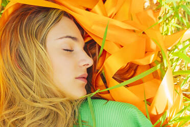 Portrait of young woman with closed eyes lying in a field with orange ribbons - ERRF03056