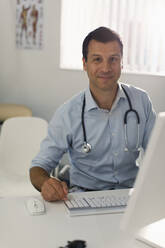 Portrait confident male doctor working at computer in doctors office - CAIF25366