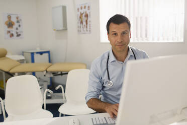 Portrait confident male doctor working at computer in doctors office - CAIF25378