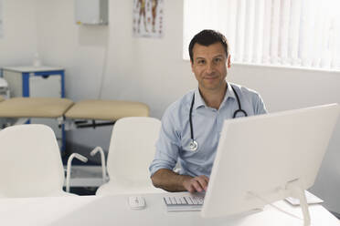 Portrait confident male doctor working at computer in doctors office - CAIF25395